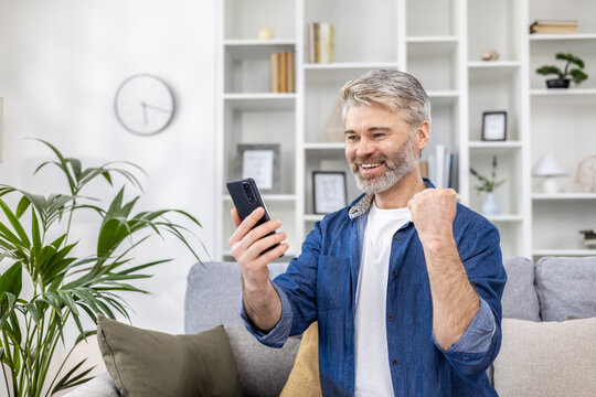 Mature gray haired man alone at home using phone and celebrating victory success happy holding hand up, adult person sitting on sofa in living room on sunny day.