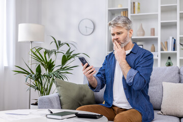 Mature adult man upset and confused reading bad news online from phone, gray haired person sitting on sofa in living room at home working with documents home finances and budget on paper work.