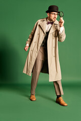 Full-length portrait of young man, detective in stylish trench coat and hat looking in magnifying...