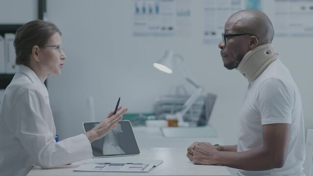 Black male patient with broken arm in cast and Caucasian female doctor sitting at desk and discussing x-ray scan on digital tablet at the consultation in clinic, side view medium shot