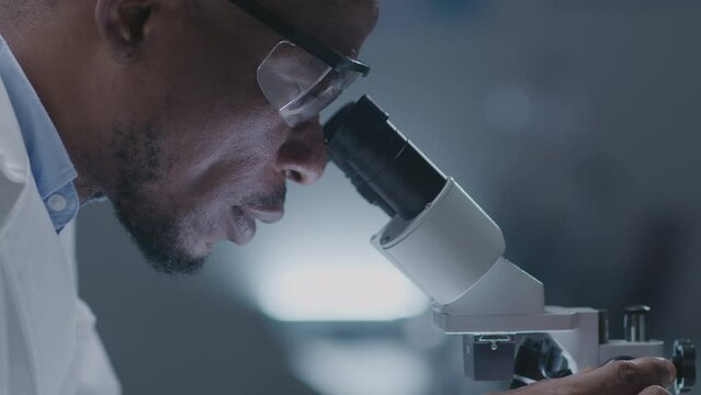 Black male scientist in lab coat looking in microscope, doing research, working with laboratory equipment. Close-up tilt-up shot