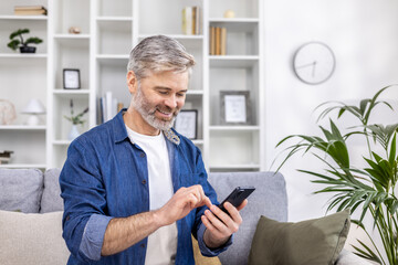 Mature adult man close-up at home smiling and using phone in living room sitting on sofa, person typing message and browsing web page.