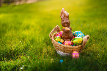 Easter eggs in basket with easter bunny on top. Chocolate rabbit with colorful decorated eggs in wicker basket in grass. Magical morning light, spring season holidays. Traditional egg hunt