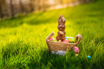 Easter eggs in basket with easter bunny on top. Chocolate rabbit with colorful decorated eggs in wicker basket in grass. Magical morning light, spring season holidays. Traditional egg hunt