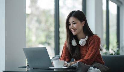 asian young woman listening music with headphone and streaming music from laptop on sofa relaxing at home