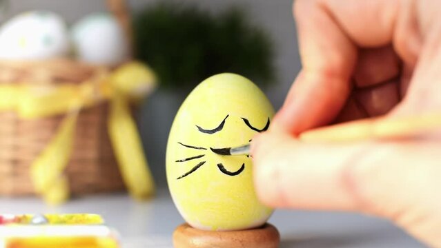 Happy Easter concept. Young womans hand draws a cute face with mustache on the eggshell of a painted yellow egg with brush and paints. Preparation for the celebration spring holiday.
