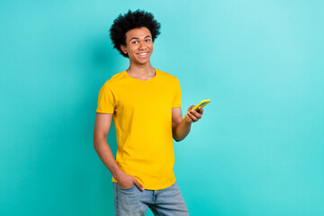 Portrait of positive funny guy afro hairdo wear yellow t-shirt hold smartphone hand in pocket isolated on shine teal color background