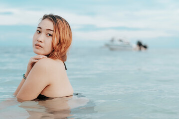 Fototapeta na wymiar A young pensive asian woman taking a dip at the beach. A female tourist unwinding in chest deep waters. Vacation scene.