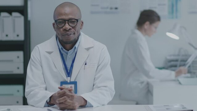 Mature African American general practitioner sitting at desk, looking at camera and smiling when working in the clinic. Medium shot