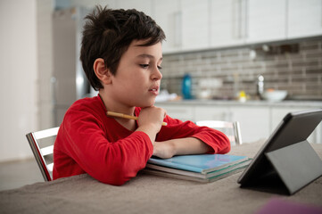 Focused Caucasian smart school boy using digital tablet at online class at home. Attentive junior...
