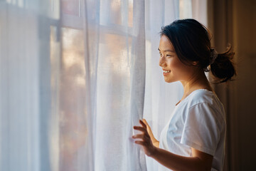 Young happy Asian woman opening curtains from her window in morning.