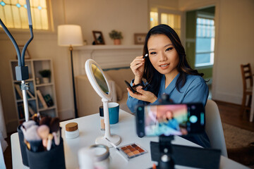 Asian makeup artist applying eyeshadow while recording beauty tutorial with smart phone.