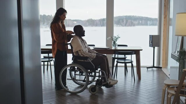 Full shot of Caucasian woman shaving head of black man in wheelchair while talking with him in living room with large picture window overlooking winter rural landscape