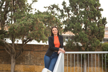 Young beautiful woman with straight brown hair, jacket and jeans, leaning on a metal railing, posing happily looking at camera. Concept fashion, beauty, trend, millennial.