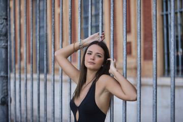 Young and beautiful woman with straight brown hair, wearing an elegant black dress, clinging to the bars of a fence with a sensual and seductive look. Concept fashion, beauty, sensuality, provocation