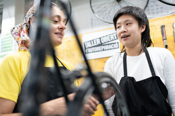 Two women in bike shop talking, a middle-aged Caucasian woman and a young Asian girl, both dressed in T-shirts and black aprons. One teaches the other bike maintenance in the workshop.