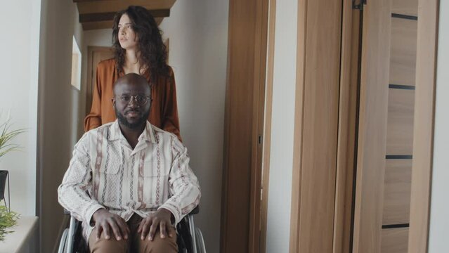Medium full shot of Caucasian woman driving black man in wheelchair along corridor towards camera. Man holding his hands on his knees and looking out window