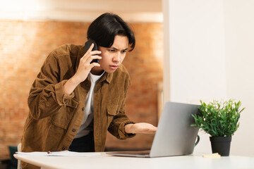 Asian Guy Talking On Phone Having Problems With Laptop Indoors