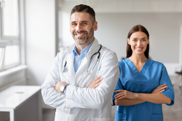 Happy male doctor and female nurse colleagues, wearing coats, standing with folded arms, posing at medical office