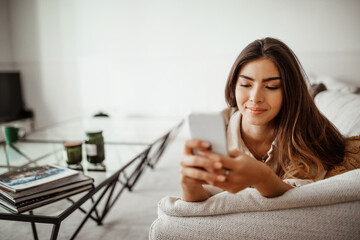 Cheerful millennial mixed race female look at phone, read message, chatting, enjoy free time, lies at sofa