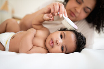 Obraz na płótnie Canvas Glad happy loving young african american woman combing hair to cute baby, lying on bed in light bedroom