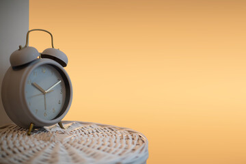 An analog large alarm clock stands on a white wicker basket a orange background.