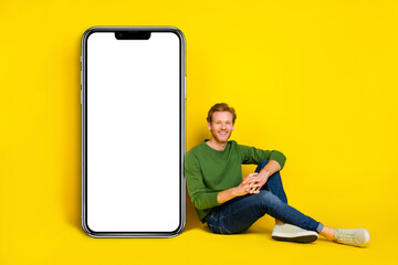 Full length photo of handsome sweet man wear pullover sitting floor modern gadget empty space isolated yellow color background