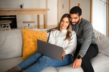 Smiling international millennial couple sit on sofa, hugging, looking at laptop, have video call in living room