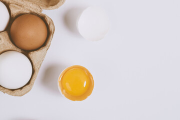 Cracked egg half with a yolk inside near the egg tray with eggs on bright background. Selective focus. Concept scene.  Top view
