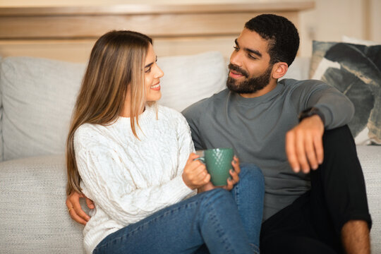 Smiling international millennial couple hugging sit on floor, enjoy free time together and coffee break with cup
