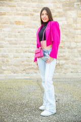 a smiling young white woman dressed in a fuchsia jacket, cropped black t-shirt and jeans, against an ancient stone wall