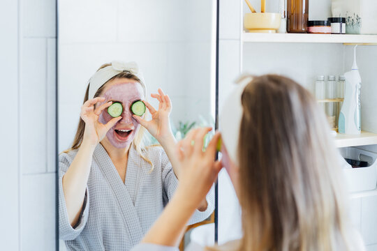 A young smiling woman with pink clay facial mask holds cucumber slices making a refreshing eye mask in bathroom. Natural cosmetic procedures for skin care at home. Beauty self-care. Selective focus