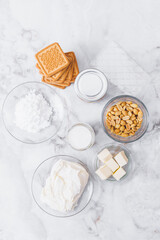 Ingredients for cheesecake. Cookies, caramel, peanuts, butter, cream cheese, powdered sugar, cream in glassware and gelatin sheets on table. Top view.