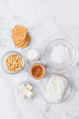 Ingredients for cheesecake. Cookies, caramel, peanuts, butter, cream cheese, powdered sugar, cream in glassware and gelatin sheets on table. Top view.