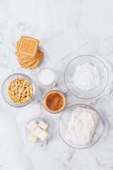 Ingredients for cheesecake. Cookies, caramel, peanuts, butter, cream cheese, powdered sugar, cream in glassware and gelatin sheets on table. Top view.