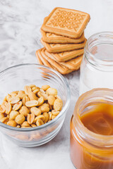 Ingredients for cooking. Stack of cookies, caramel, heap of peanuts, cream in glassware on table. Selective focus.