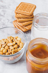 Ingredients for cooking. Stack of cookies, caramel, heap of peanuts, cream in glassware on table. Selective focus.