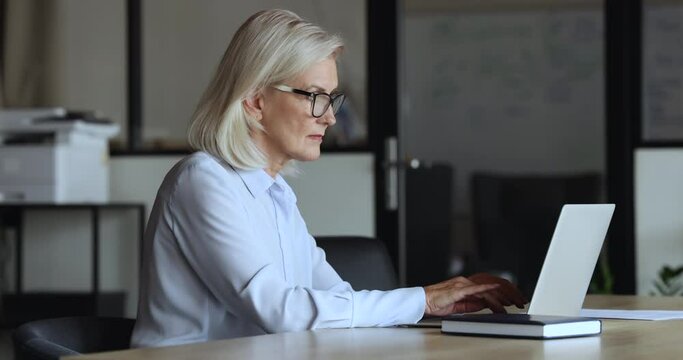 Side view serious aged businesswoman in glasses working on laptop, typing message, make business remotely, consulting to client through e-mail sit at workplace desk. Workflow use modern wireless tech