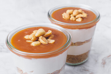 Cheesecake in a jar. Layers of grinded cookies, caramel, peanuts and  cream cheese. Covered in caramel and decorated with peanuts. Close-up photo. Selective focus. Macro shot.