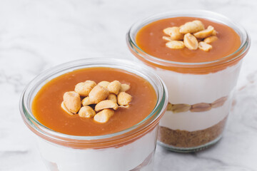 Cheesecake in a jar. Layers of grinded cookies, caramel, peanuts and  cream cheese. Covered in caramel and decorated with peanuts. Close-up photo. Selective focus. Macro shot.