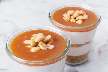 Cheesecake in a jar. Layers of grinded cookies, caramel, peanuts and  cream cheese. Covered in caramel and decorated with peanuts. Close-up photo. Selective focus. Macro shot.