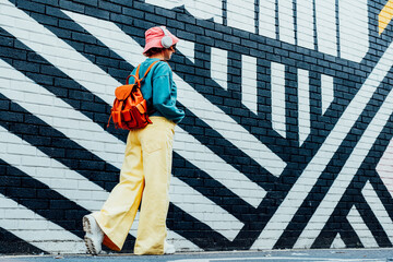 Back view stylish woman in bright clothes and bucket hat wearing wireless headphones and listening...