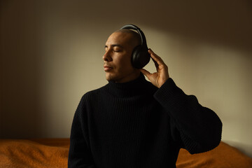 A caucasian man sitting on a bed wearing wireless headphones listening. 