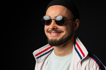 Obraz premium A young, tough guy from the streets dressed in a tracksuit and round sunglasses, posing, isolated on a black background. Urban street culture, Street gang, urban fashion.