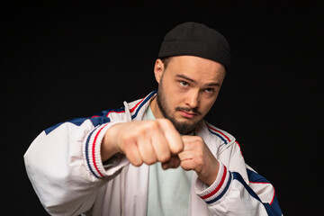 A young, tough guy from the streets dressed in a tracksuit, has clenched his fists and is ready to strike. Isolated on a black background. Urban street culture.