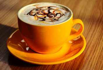 Cappuccino in isolated yellow ceramic cup. decorative froth on top with caramel and brown chocolate line drops. brown wood pattern background. gourmet coffee drink concept. 