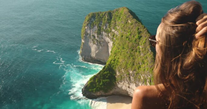 Tourist girl enjoy sea view standing on the mountain top. Young woman back view relax standing over Tyrannosaur rock at Kelingking Beach, Nusa Penida, Indonesia. Famous touristic place. Slow Motion 4K
