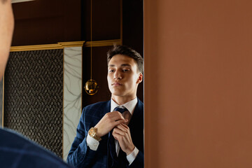 The young man in a formal dark blue suit and golden watches adjusts his tie in front of the mirror.