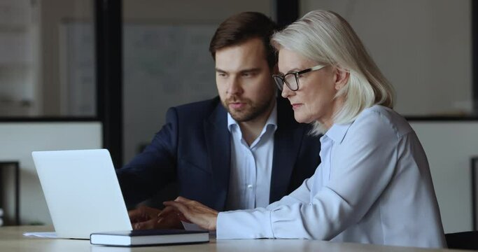 Older and younger office employees working on-line on laptop, look at laptop screen, discuss new task, corporate software, review new business apps, share ideas, engaged in teamwork use modern tech