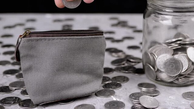 Woman Putting Some Cents into a Small Change Grey Purse. Scattered silver Ukrainian coins, change, pennies, glass piggy bank lie on the blurred background of a table. Savings, accumulation. Poverty.